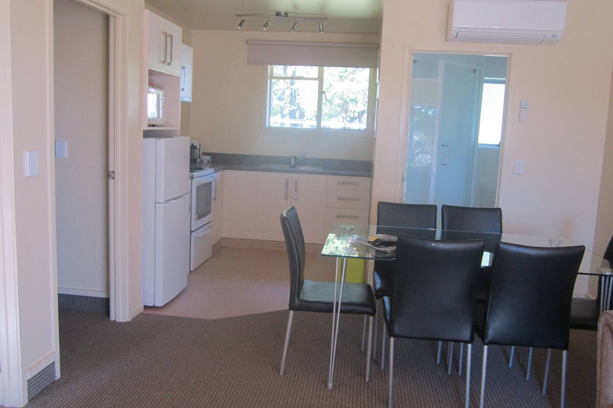 A compact kitchen with white appliances, a dining area with a glass table and black chairs, and a view through a window.