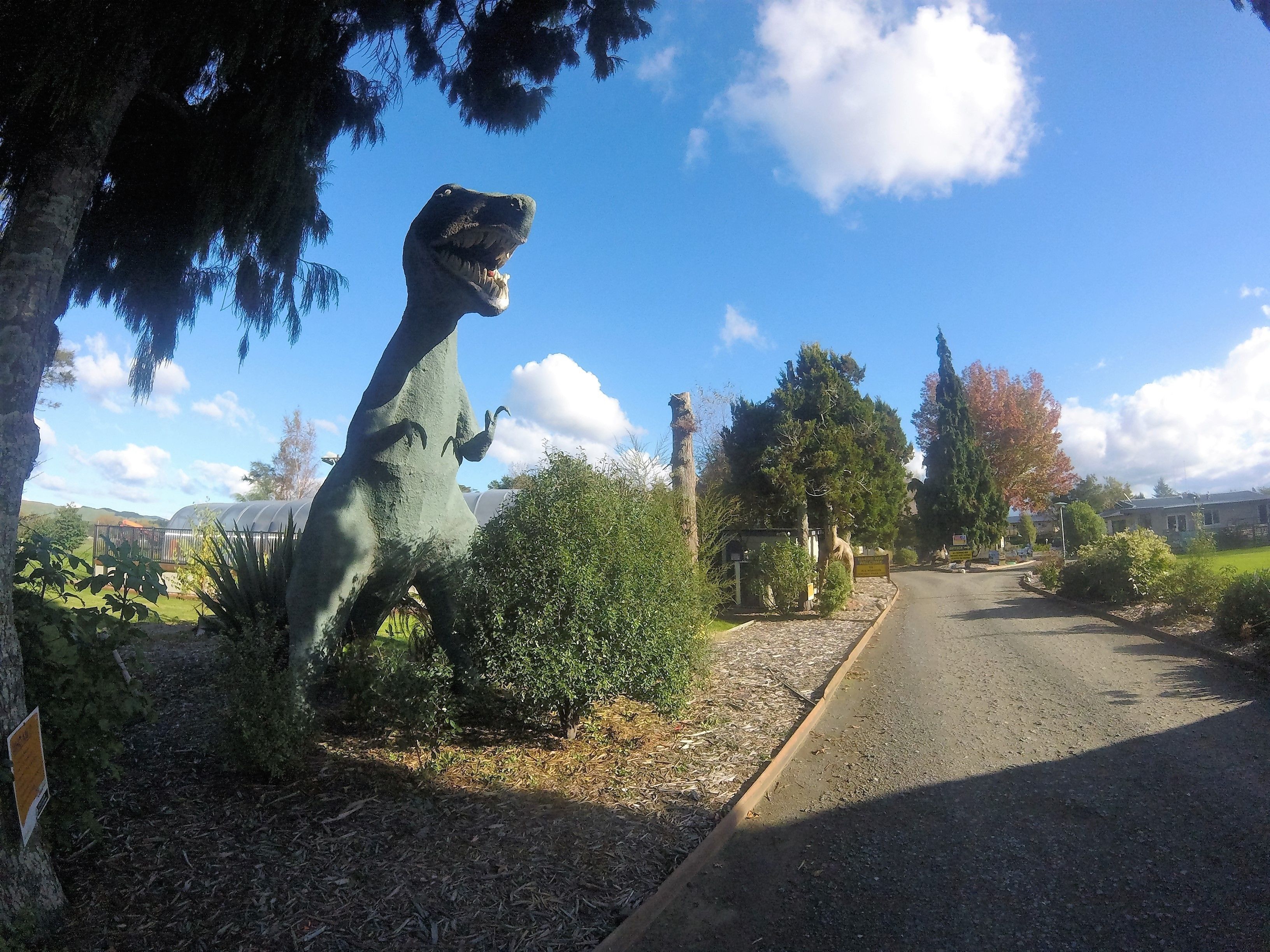 Uma estátua de dinossauro em um parque, com uma estrada, árvores e céu claro.