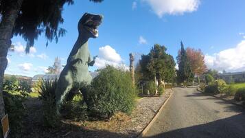 Uma estátua de dinossauro em um parque, com uma estrada, árvores e céu claro.