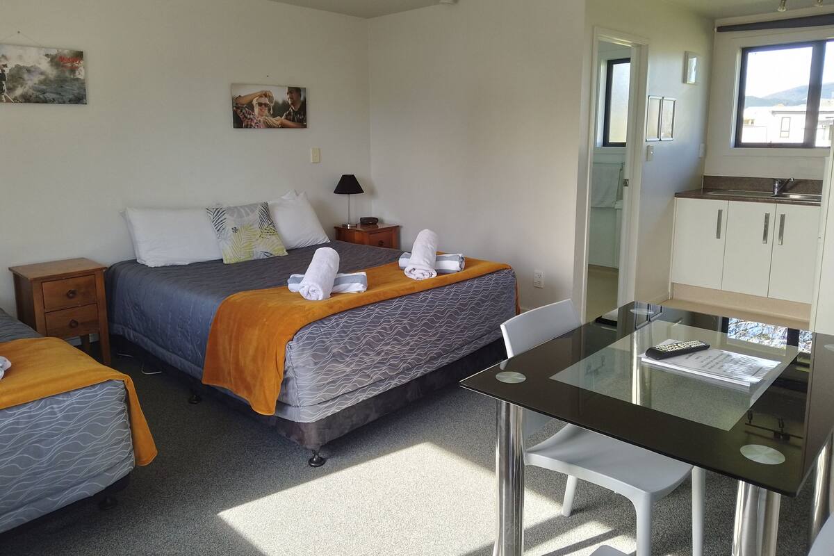 A hotel room with a bed, a small table, a chair, a nightstand, a lamp, and a kitchen area.