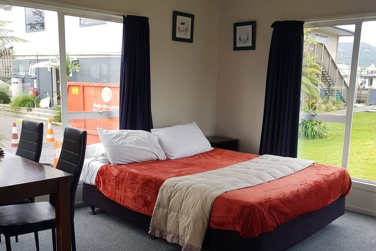 A single bed with a red and beige bedspread, a small table with two chairs, and a window with curtains.