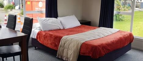 A single bed with a red and beige bedspread, a small table with two chairs, and a window with curtains.