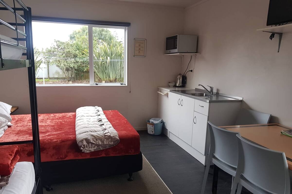 A dormitory room with a bunk bed, a desk, a chair, a microwave, and a window with a view of greenery.