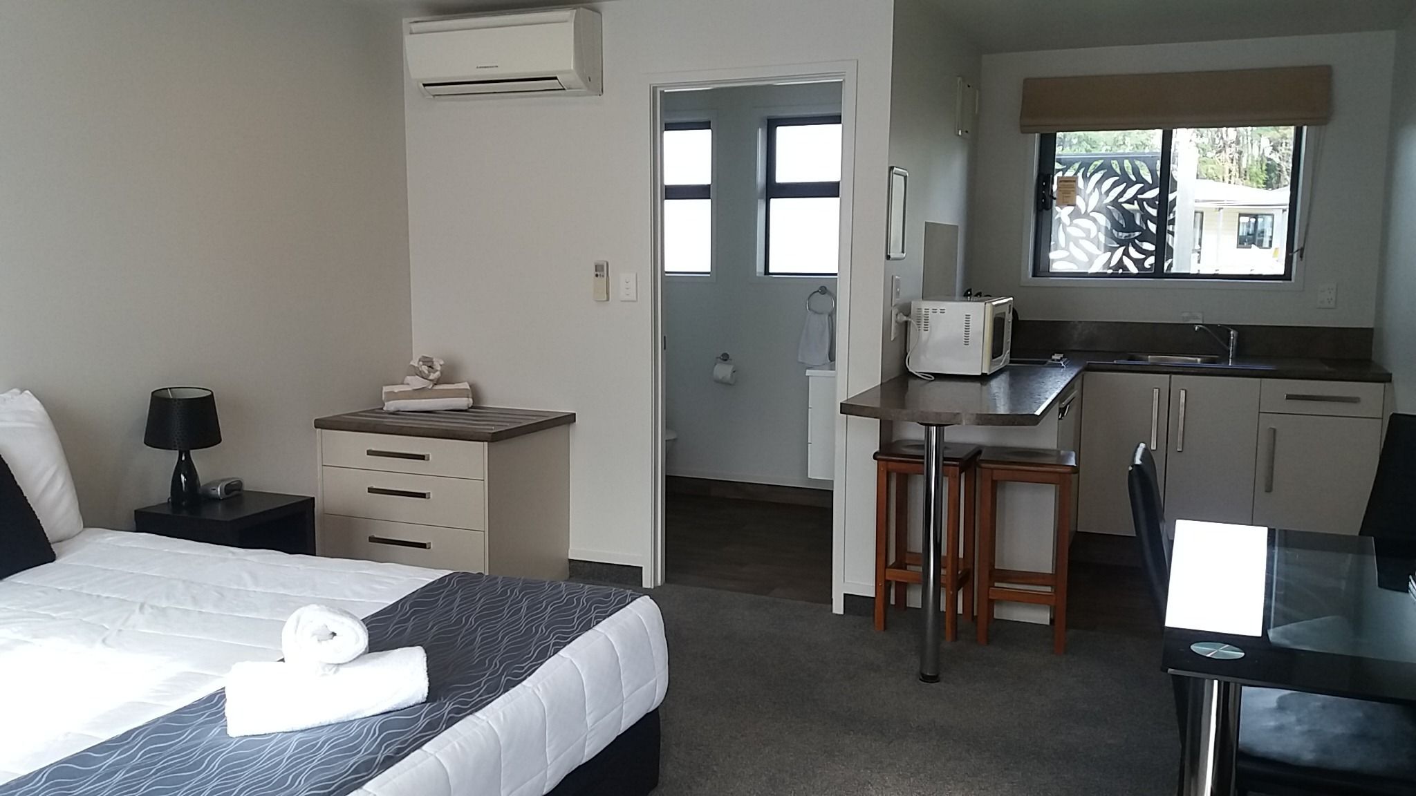 Een compacte hotelkamer met een bed, nachtkastje, lamp en een kitchenette met een magnetron en barstoelen.