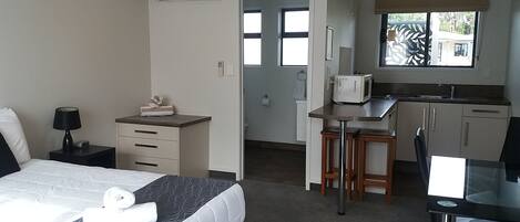 Een compacte hotelkamer met een bed, nachtkastje, lamp en een kitchenette met een magnetron en barstoelen.