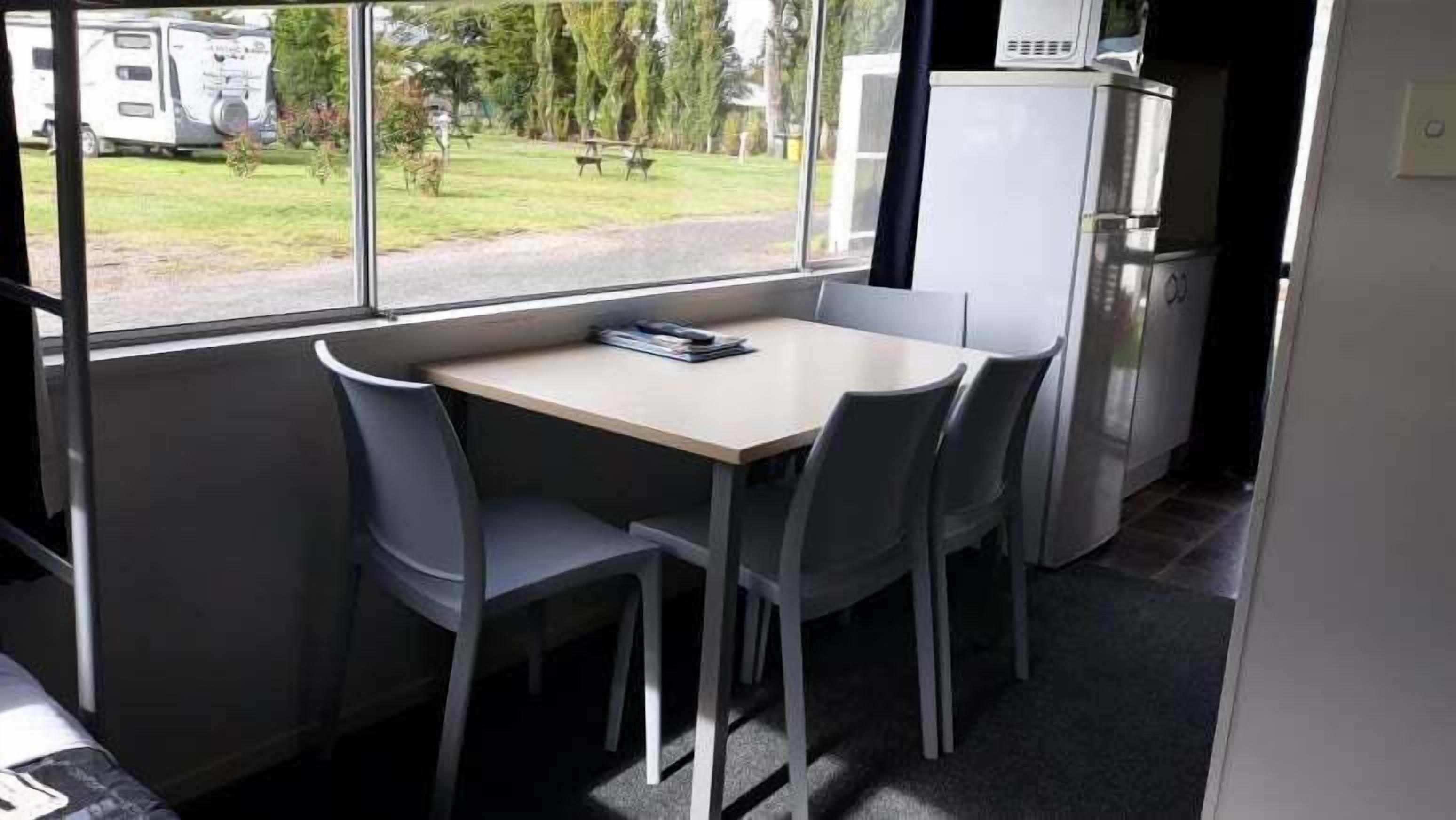 A compact kitchenette with a refrigerator, microwave, and a small table with chairs. A window reveals a grassy outdoor area with trees and a picnic table.