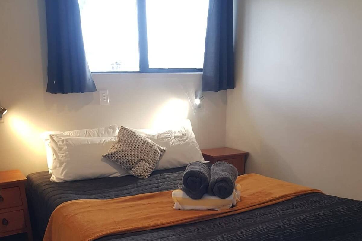A neatly made bed with a dark blue bedspread, a white pillow, and a rolled towel on top. There is a small wooden nightstand beside the bed with a lamp on it.
