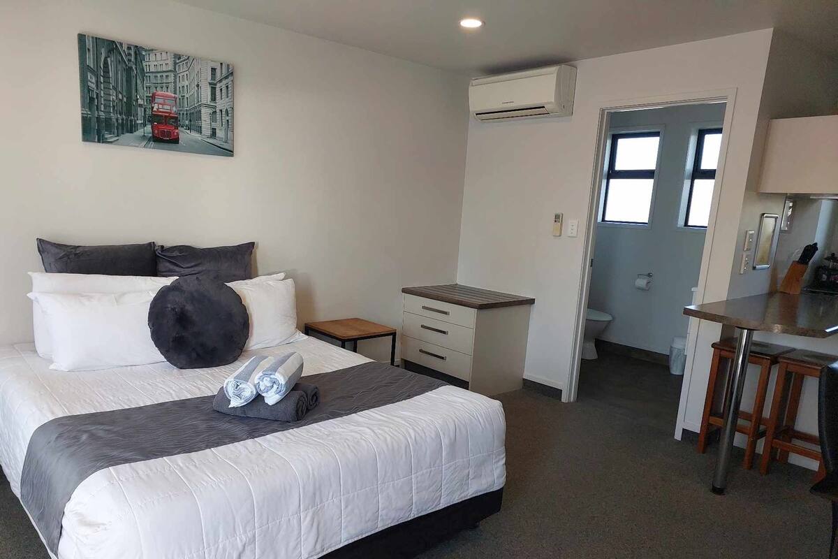 A hotel room with a bed, a small bedside table, a desk with a chair, an air conditioning unit, and a bathroom visible through an open door.