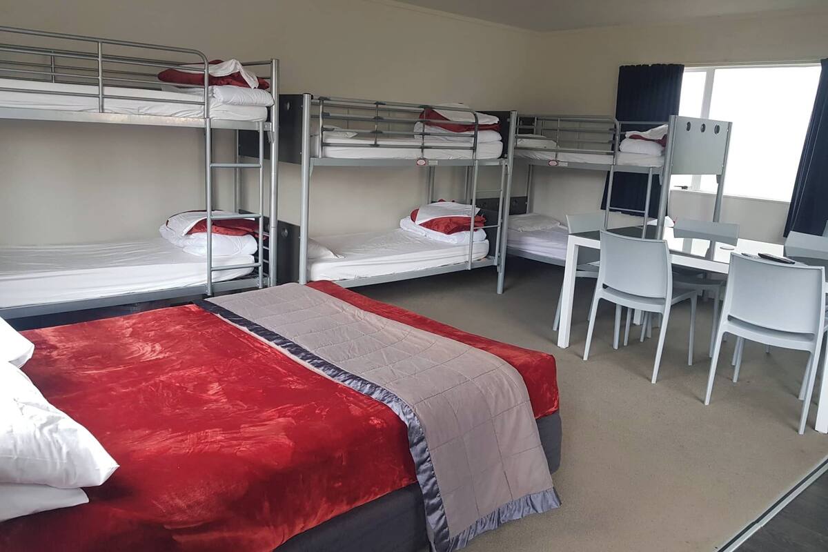 A dormitory room with bunk beds, a desk, chairs, and a bed with a red blanket.