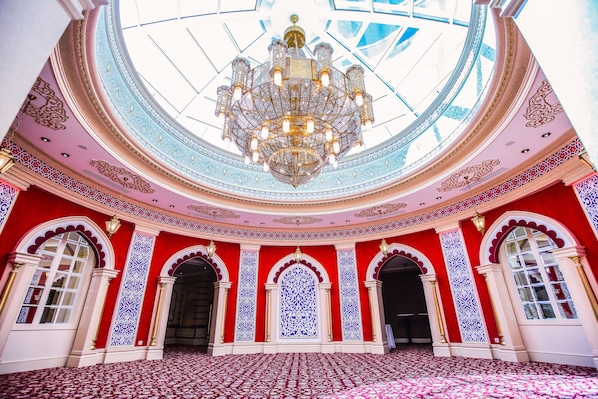 Un salón majestuoso con una gran araña, paredes con estampado azul y rojo, y un techo abovedado.