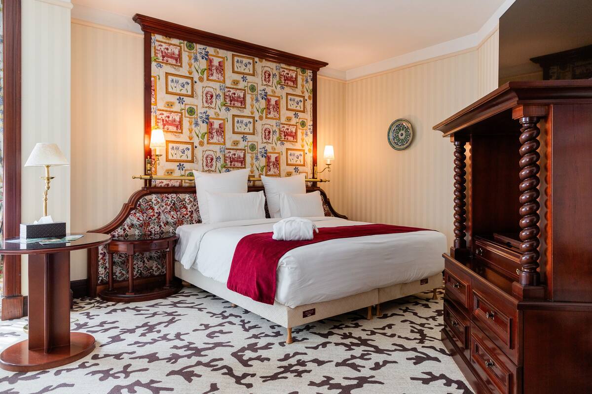 A hotel room with a large bed, a wooden dresser, a round table, and a patterned wallpaper.