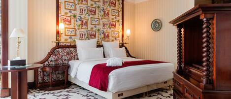 A neatly made bed with a red blanket, a wooden headboard, and a patterned wallpaper. There is a wooden dresser to the right and a small table with a lamp on the left.