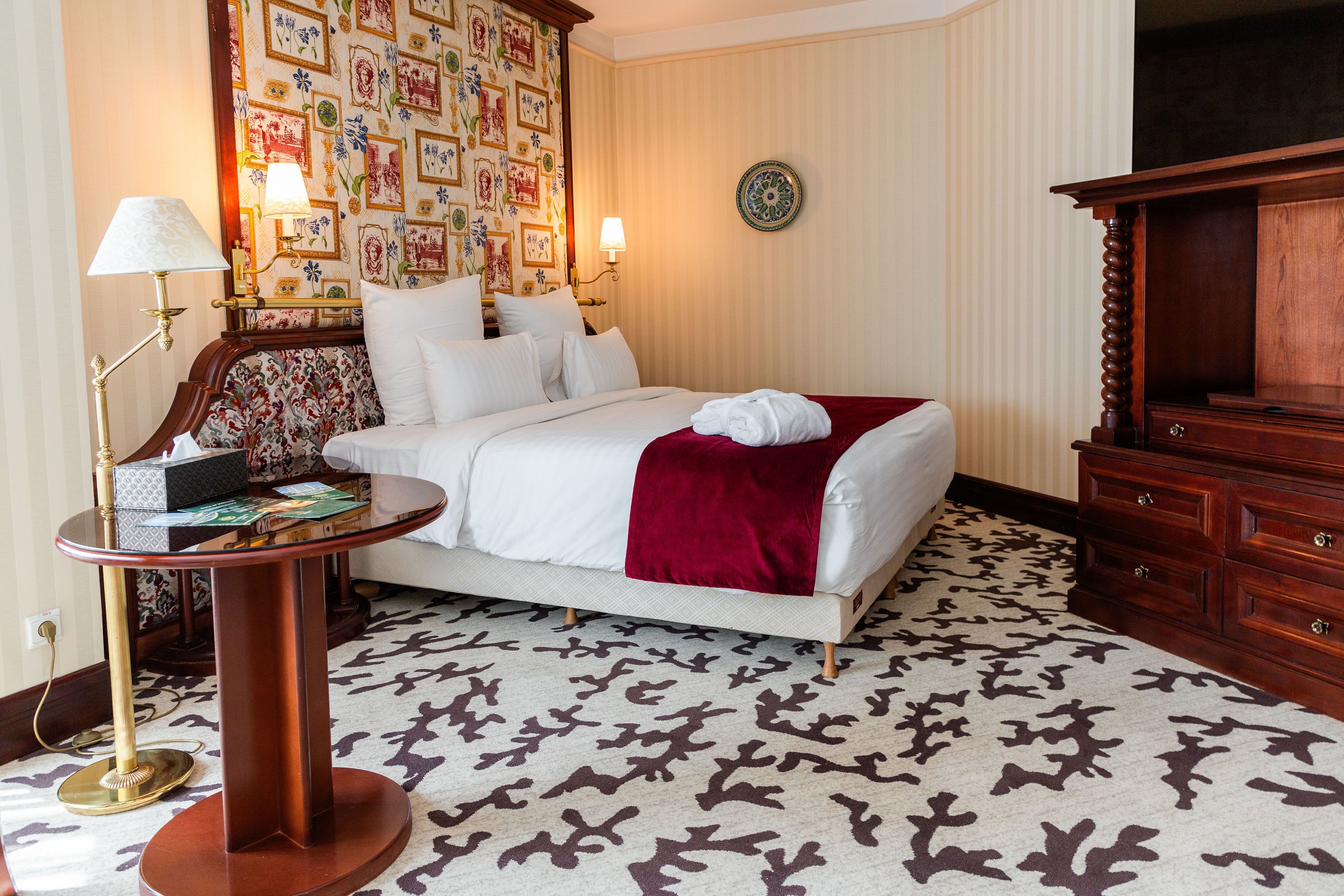 Habitación de hotel con una cama, una mesita de noche con lámpara, un muro decorativo, un televisor y una alfombra estampada.