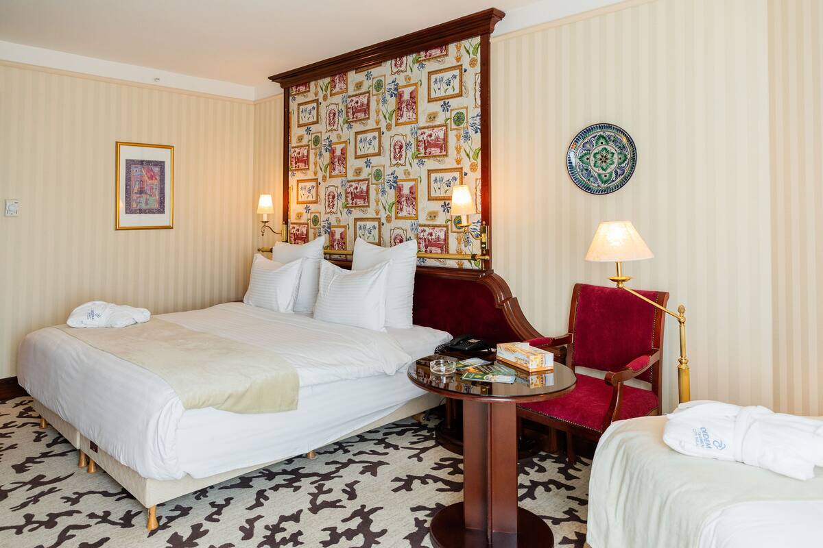 A hotel room with a large bed, a red armchair, a small table with a lamp, and a decorative wall panel.