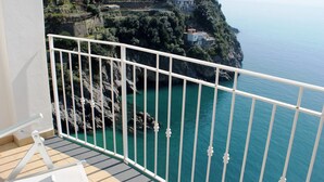 A balcony with a white railing overlooking a coastal landscape with terraced vineyards and a clear blue sea.