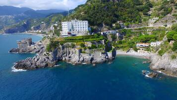 Un hotel costero con ubicación en un acantilado, que cuenta con un edificio grande con múltiples balcones, una playa y una fortaleza histórica cercana.