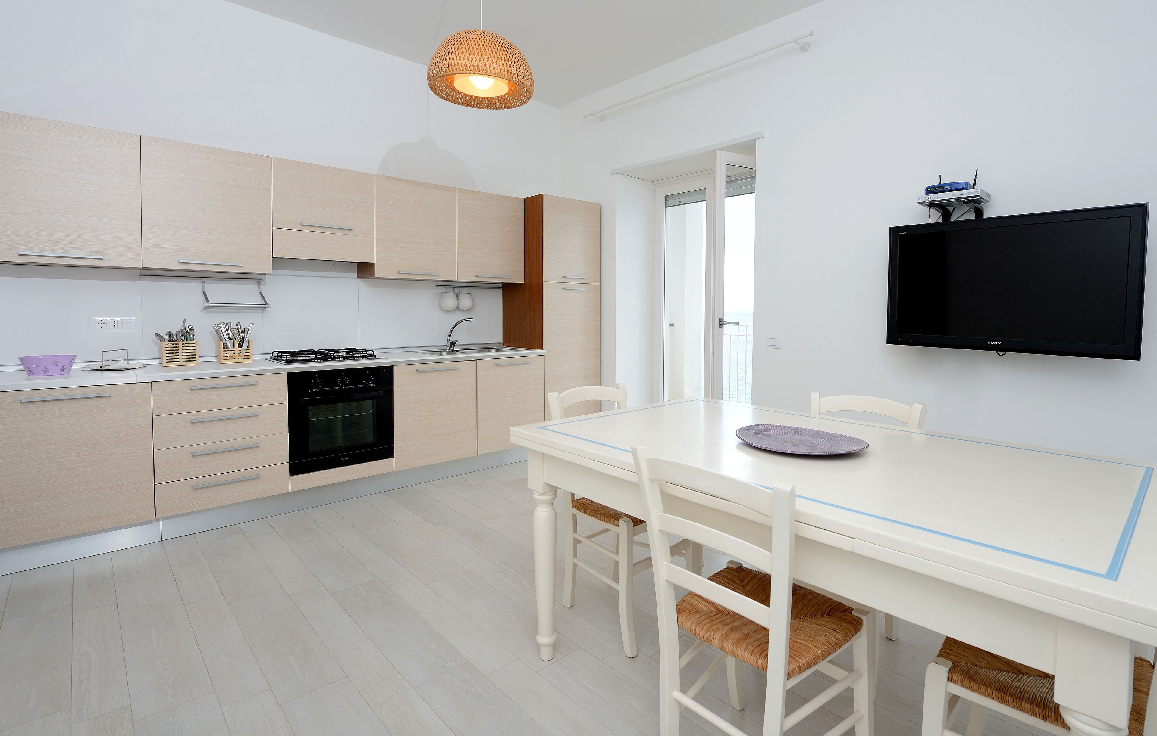 Une cuisine moderne avec des meubles en bois clair, une table à manger blanche et un téléviseur à écran plat fixé au mur.