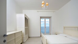 A bedroom with a bed, a wardrobe, a chest of drawers, a nightstand, and a window with a view of the sea.