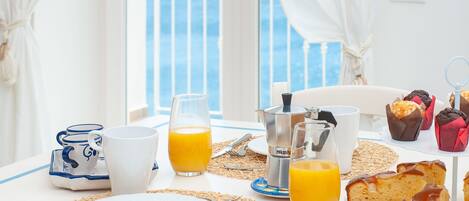 Una mesa de desayuno con mantel blanco, una cafetera, un vaso de zumo de naranja, un plato y muffins.