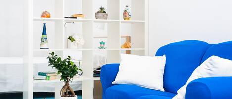 A modern living room with a blue sofa, white cushions, a white coffee table, and a white bookshelf with various decorative items.