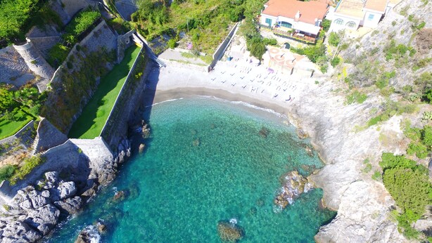 A coastal area with a clear beach, rocky cliffs, and a building with a red roof.
