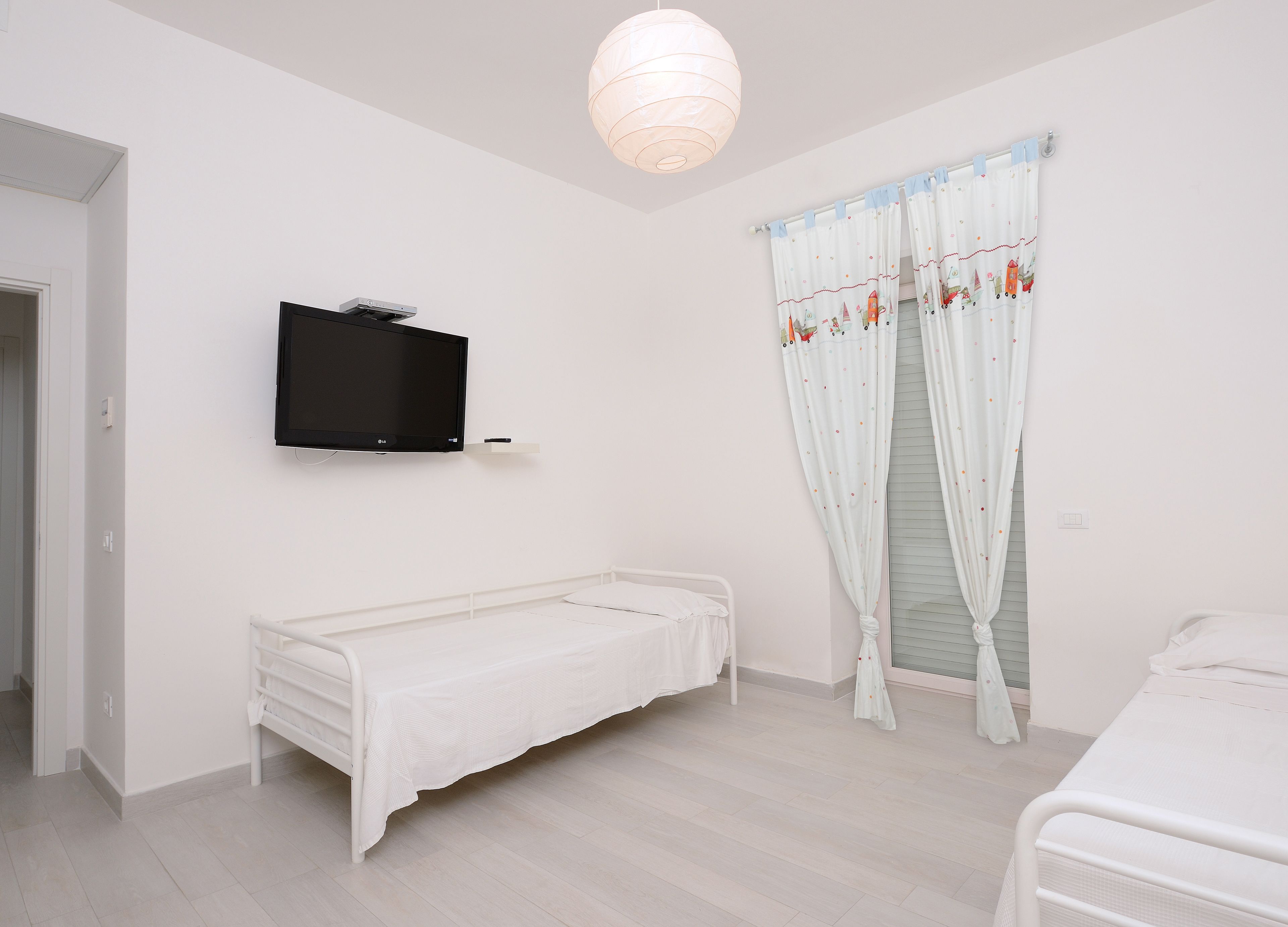 A bedroom with a single bed, a television mounted on the wall, and a window with curtains.
