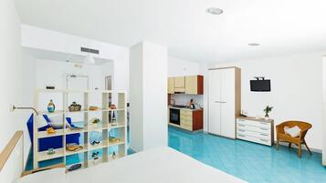 Une pièce moderne et bien éclairée, comprenant un lit, une armoire blanche, un petit coin cuisine et un espace salon avec un fauteuil.