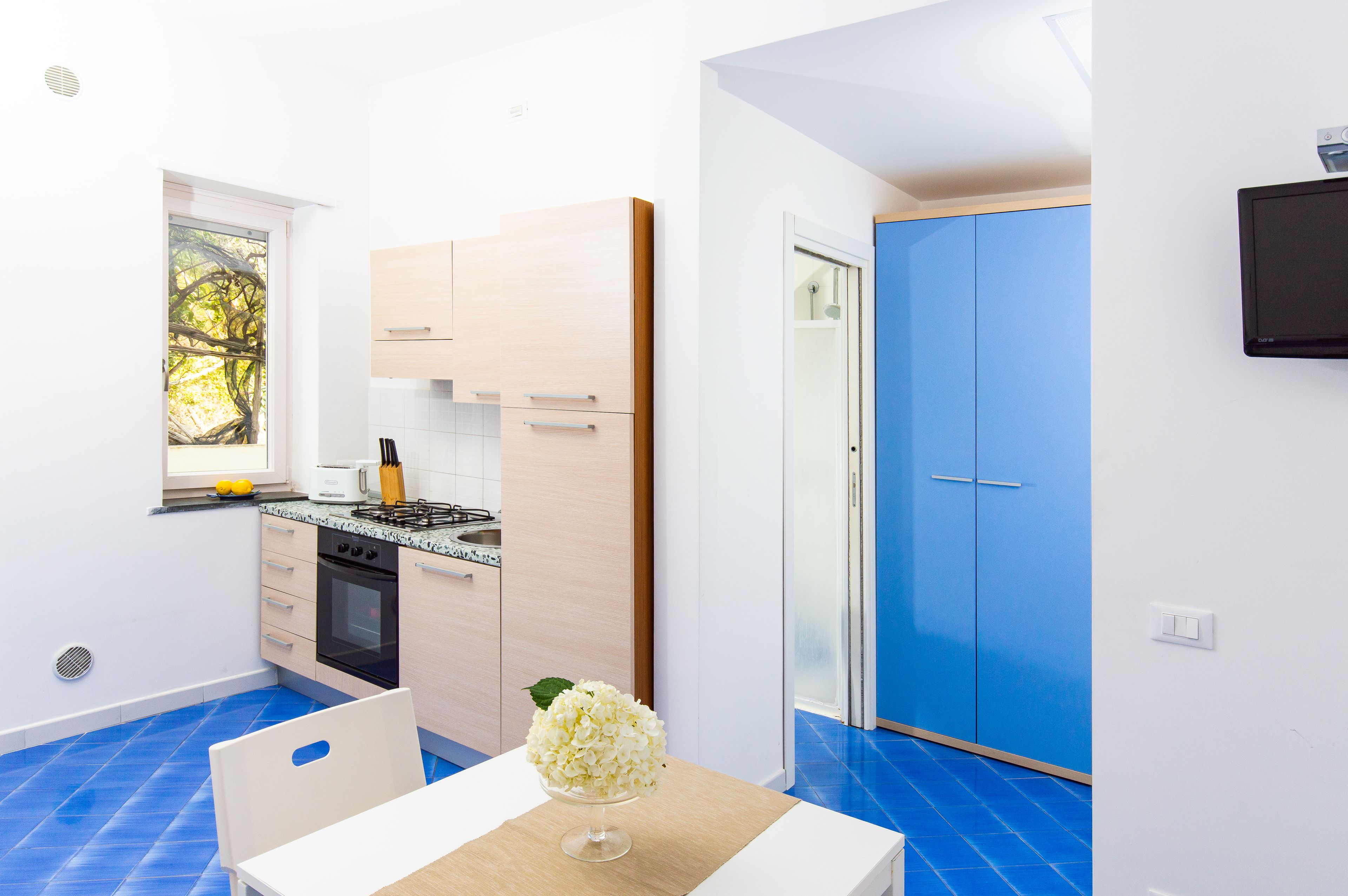 A compact kitchen with a stove, oven, and a sink. There is a dining table with a chair and a vase of flowers. A blue door and a window with a view of trees are visible.