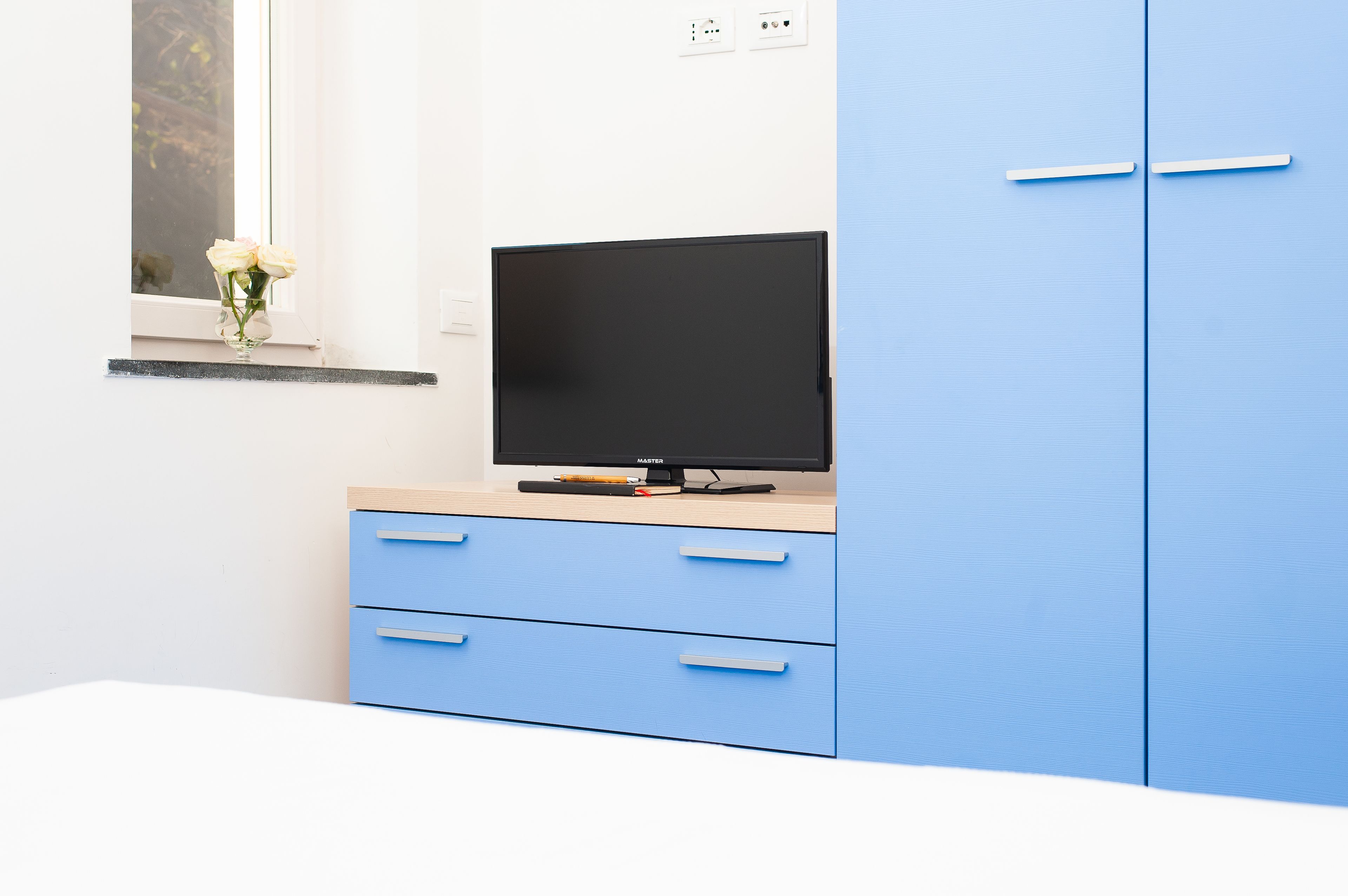 A room with a blue cabinet, a television on top, and a window with a vase of flowers.