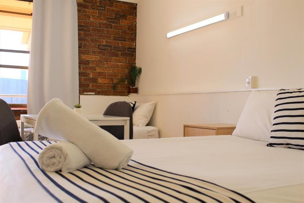 Habitación de hotel con cama, escritorio, silla, mesita de noche, lámpara y pared de ladrillo.