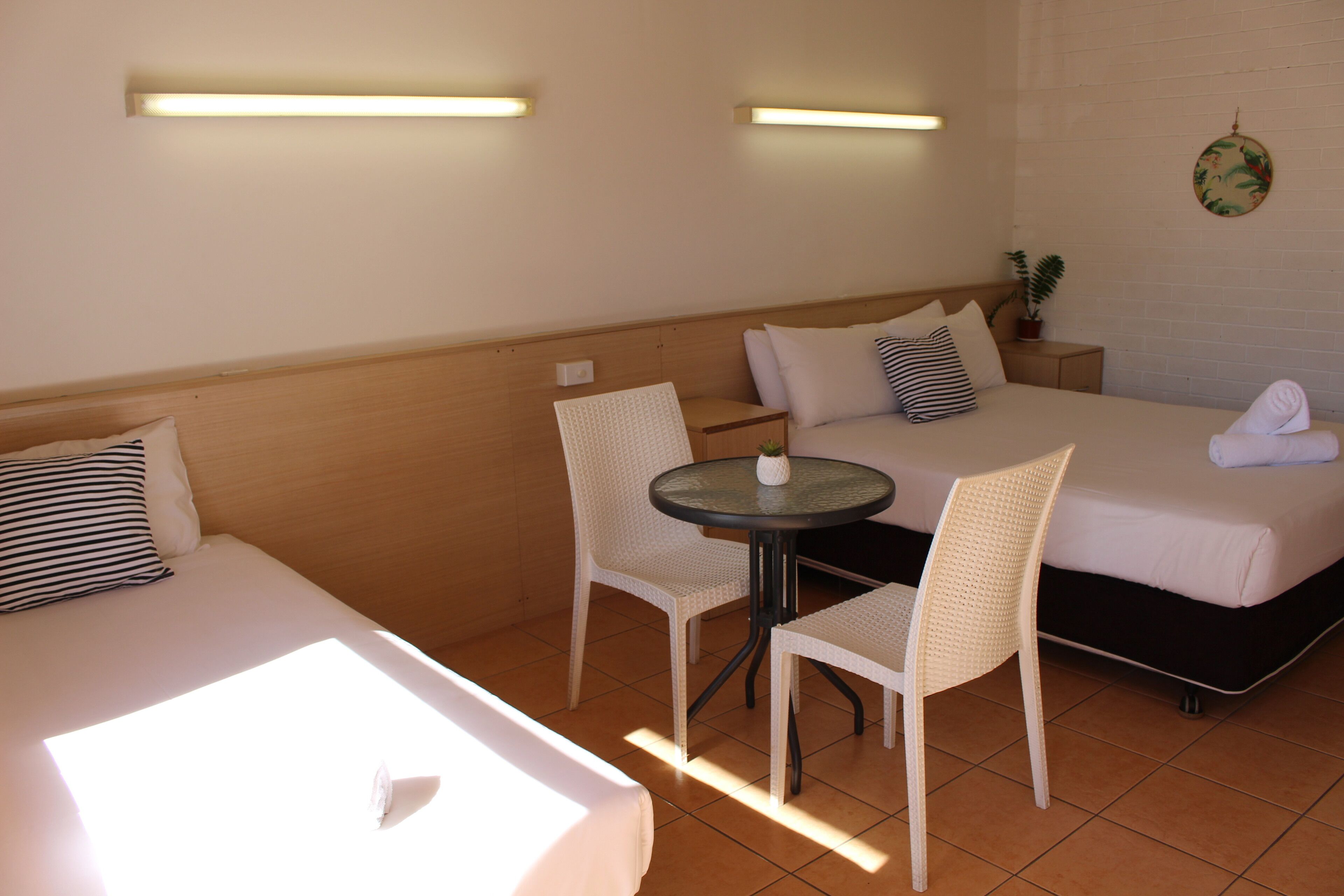 A hotel room with two beds, a round table, and chairs. There are wall-mounted lights and a potted plant.