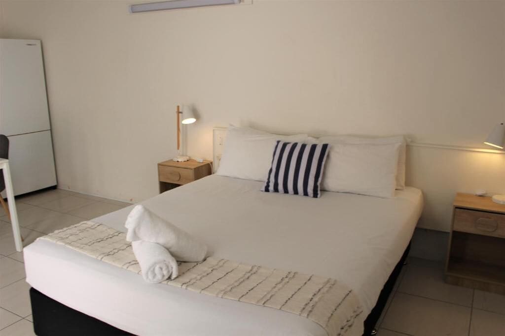 A neatly made bed with white linens and striped pillows, a bedside lamp, and a small wooden nightstand.