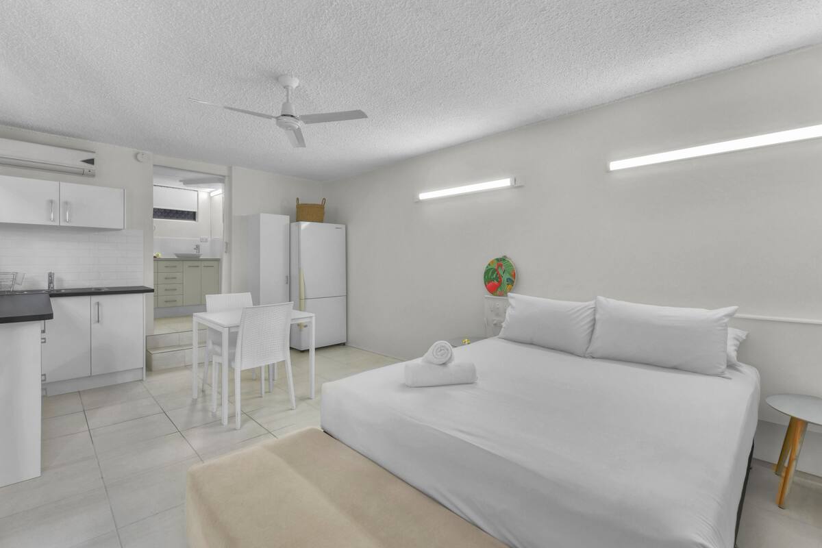 A modern hotel room with a bed, a desk, a chair, a refrigerator, and a ceiling fan.