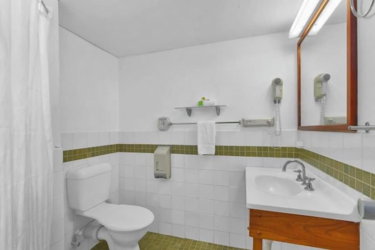 Un baño con inodoro, lavamanos y espejo. Hay secadores de pelo en la pared.