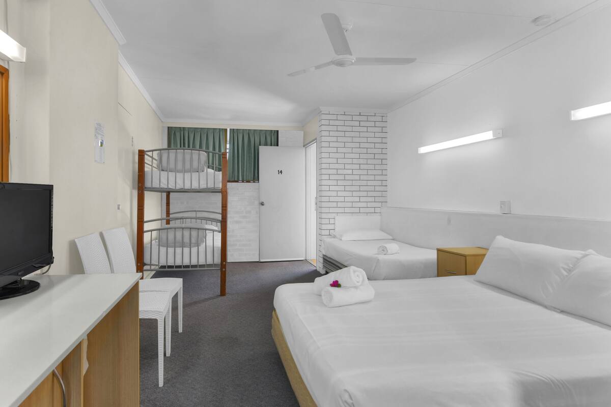 A hotel room with bunk beds, a desk with a computer, a bed with a rolled towel, and a white brick accent wall.
