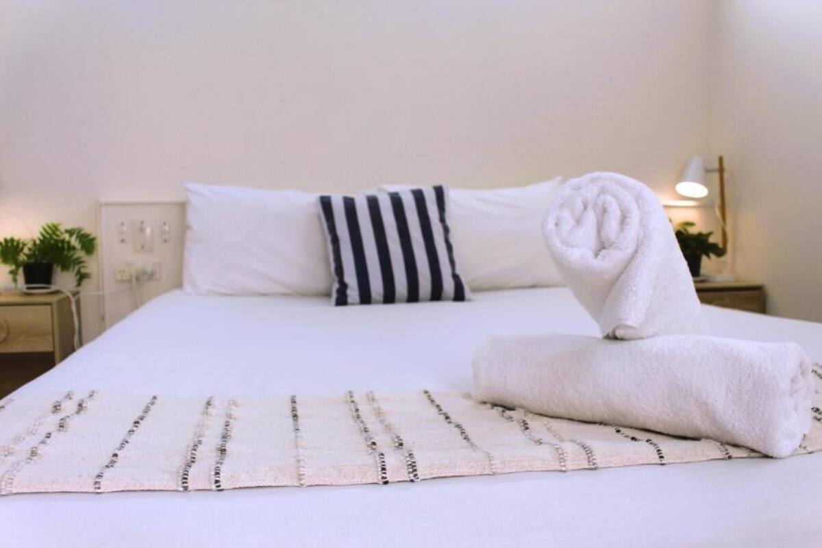 A neatly made bed with white linens, a striped pillow, and a rolled towel on a beige rug.