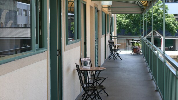 A covered outdoor seating area with tables and chairs, a railing, and a view of trees and buildings in the background. - Astray Motel (Rotorua)