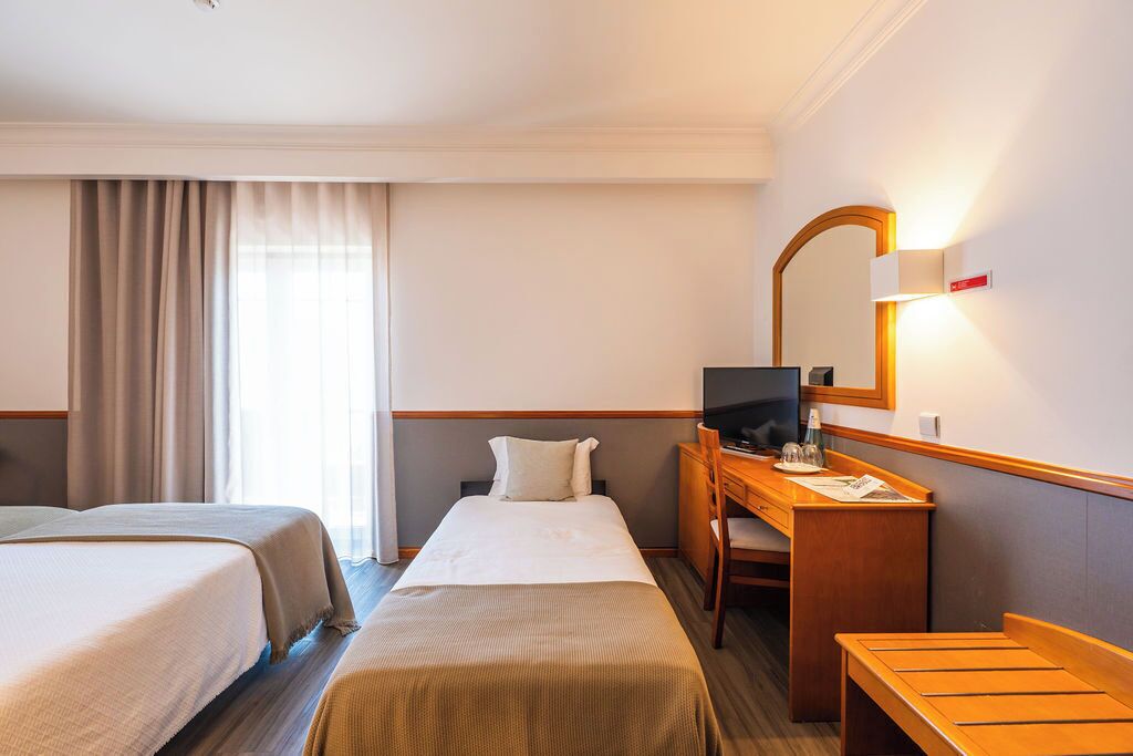 A hotel room with two beds, a desk with a computer, a chair, and a bench.