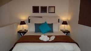 Una cama bien hecha con edredón blanco y manta marrón. Hay dos lámparas de noche a cada lado de la cama, y un par de zapatillas colocadas sobre ella.