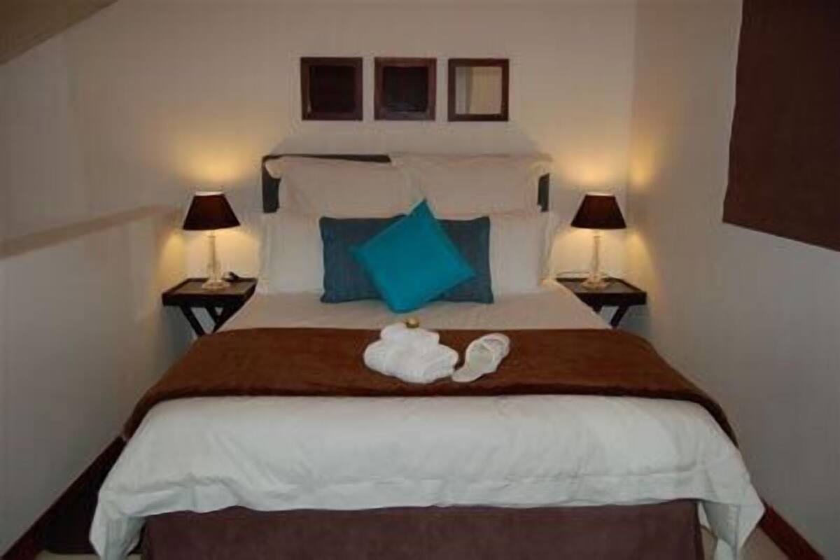 Un lit bien fait, recouvert d’une couette blanche et d’un plaid marron. De chaque côté du lit, on trouve une lampe de chevet, et une paire de chaussons est posée sur le lit.