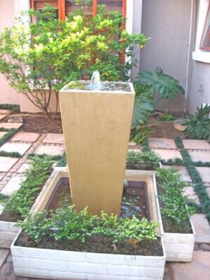 Un élément décoratif aquatique rectangulaire, doté d’une fontaine centrale, entouré d’un bac carré rempli de plantes vertes.