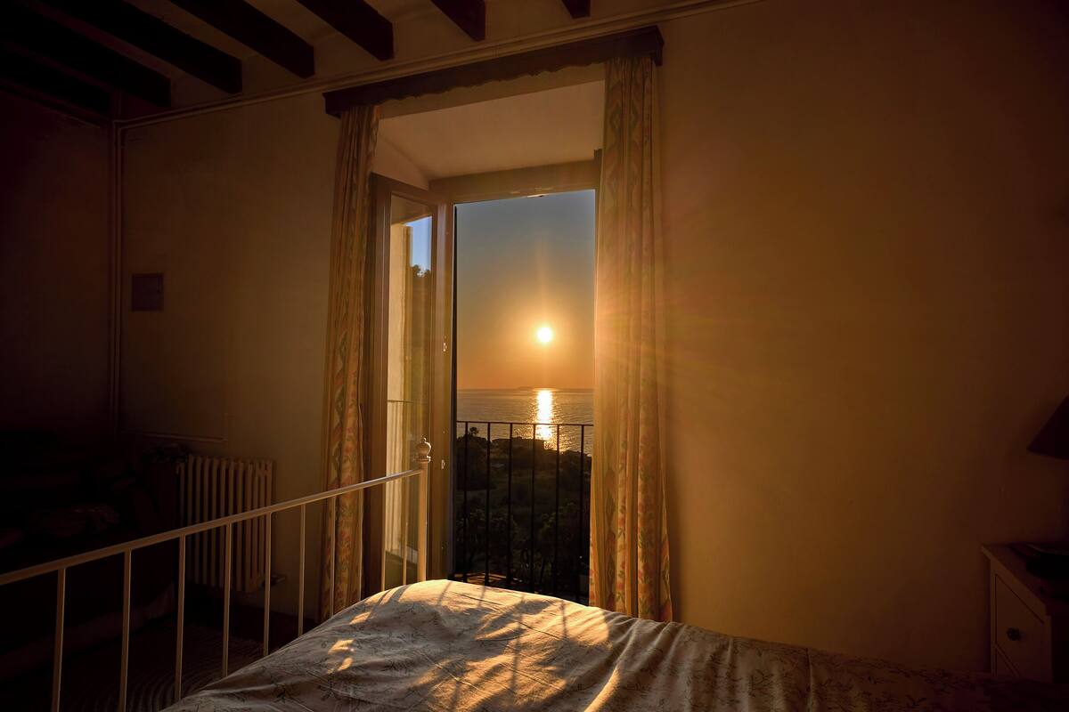 A bedroom with a balcony, a bed, and a view of the sunset over the water.