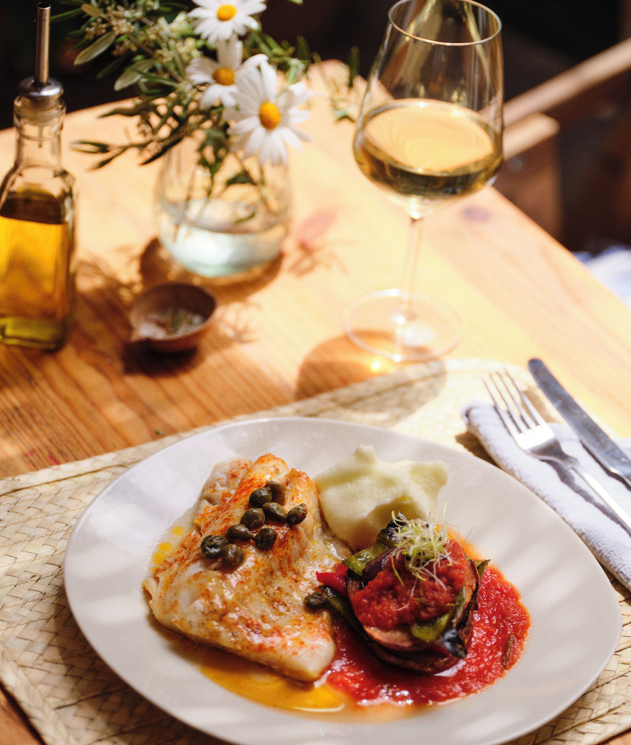 Une assiette de nourriture contenant du poisson, des légumes et un verre de vin blanc, posée sur une table recouverte d’un set de table.