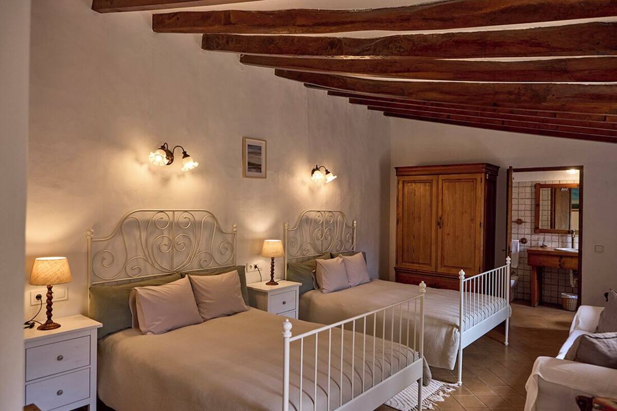 A bedroom with two beds, wooden beams on the ceiling, a wooden wardrobe, and a bathroom visible in the background.