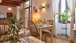 Ein Essbereich mit Holzkästen und einem Tisch, gedeckt für das Frühstück. Ein Fenster mit Vorhängen, eine Pflanze auf einem Beistelltisch und eine Wandleuchte sind zu sehen.