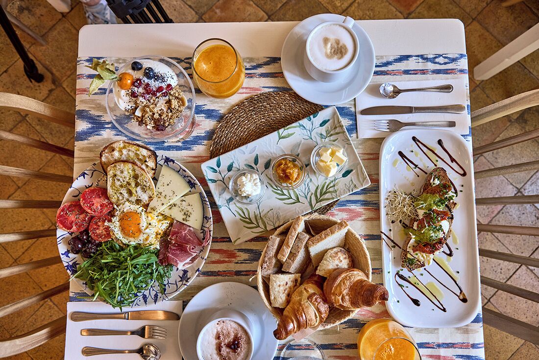 A breakfast setting with various dishes, including eggs, bread, and a croissant, on a table with a patterned tablecloth.