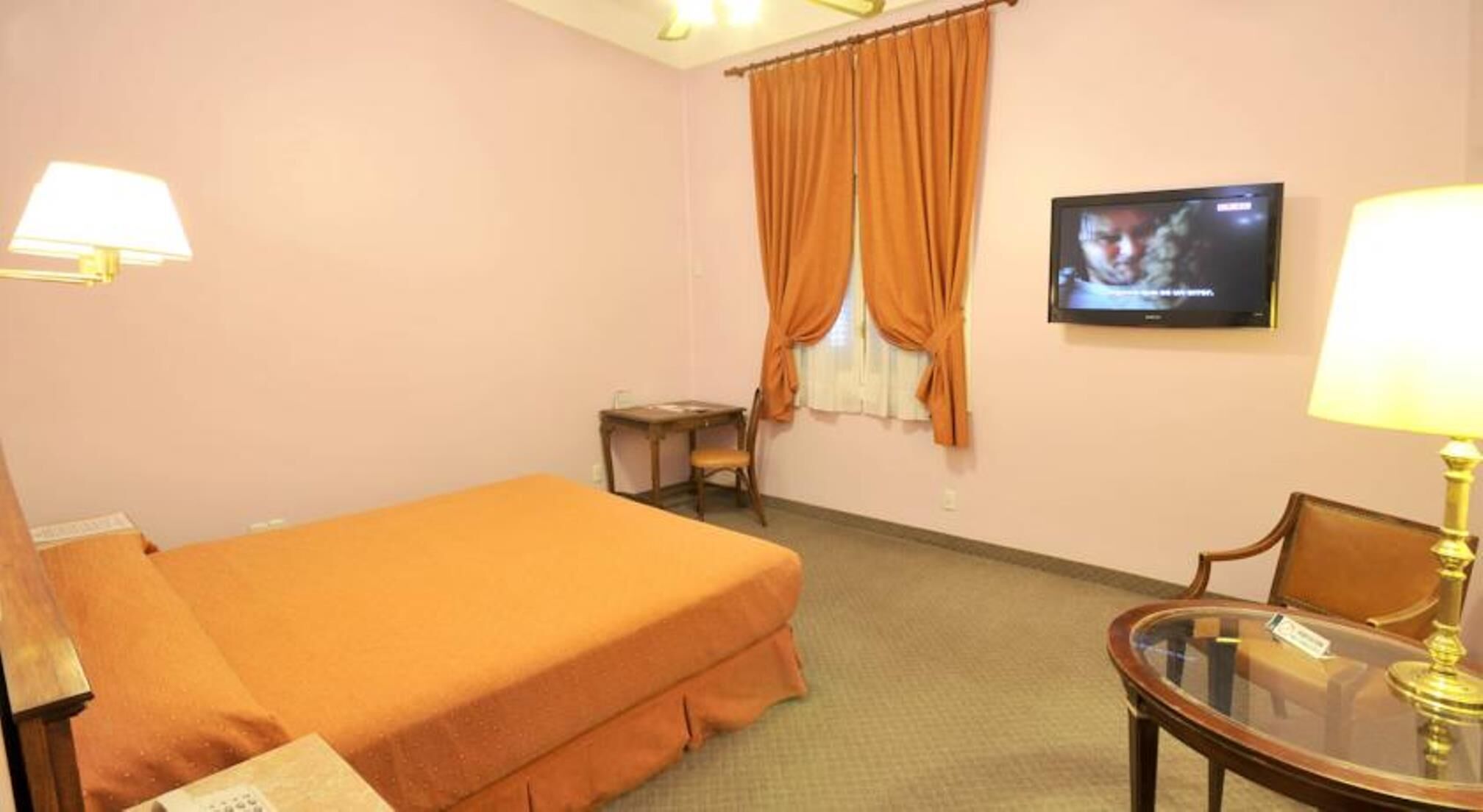 A hotel room with a single bed, a small table, a chair, a television, and a lamp.