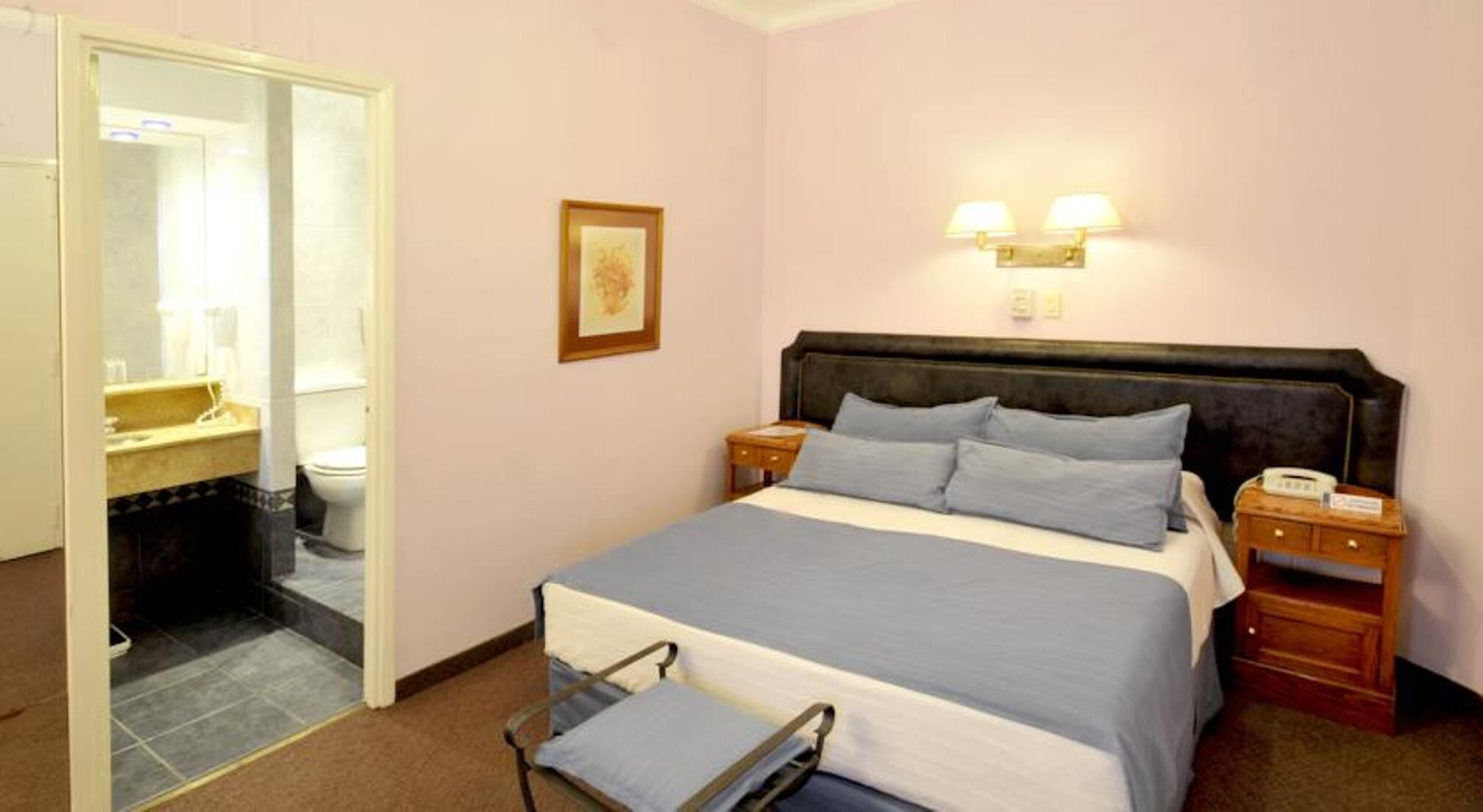 Een hotelkamer met een bed, nachtkastjes, een stoel, een spiegel en een zichtbare badkamer door een geopende deur.
