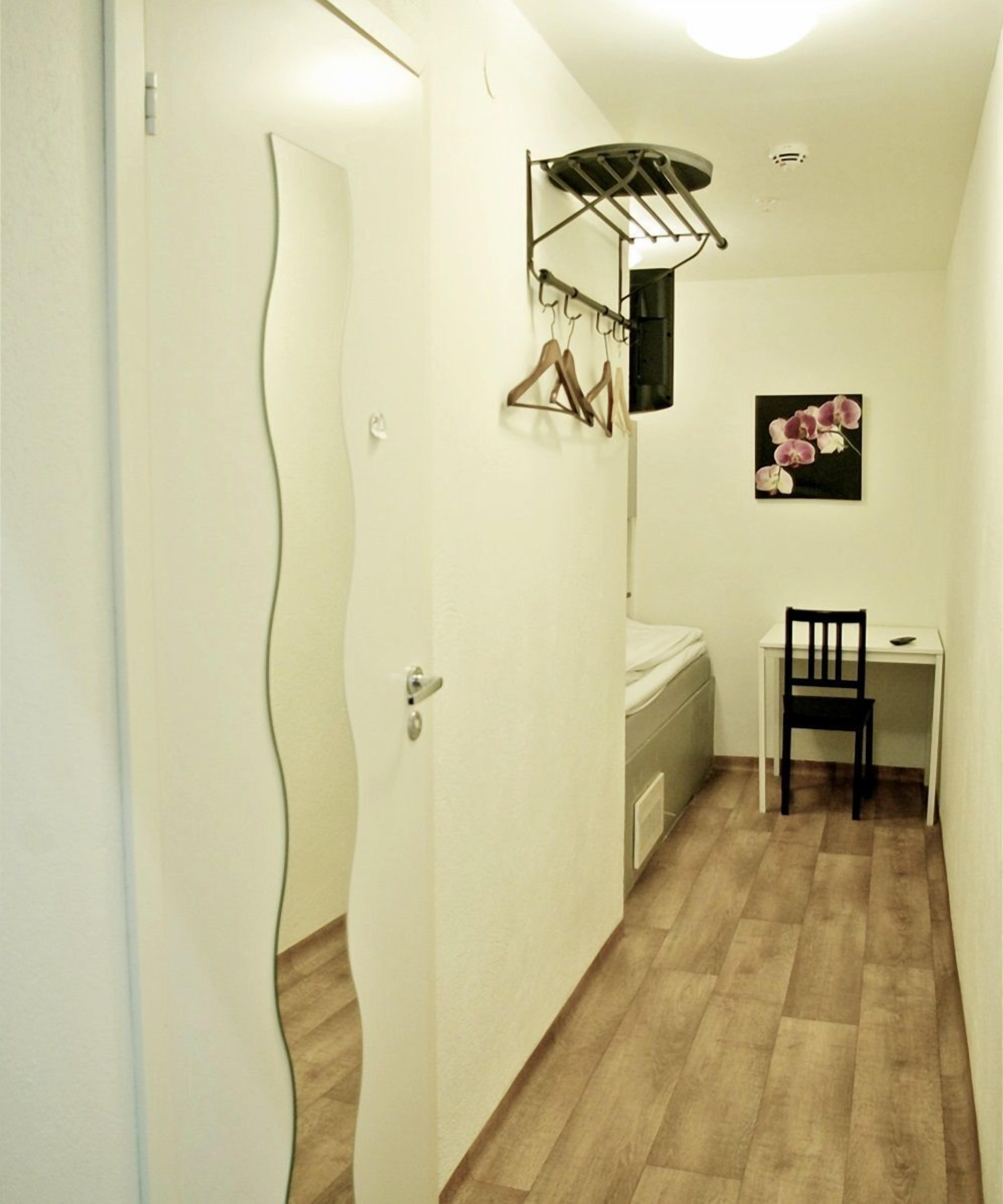 Un pasillo estrecho con suelo de madera, una puerta blanca, un escritorio pequeño con una silla, un radiador y un tendedero con ropa.