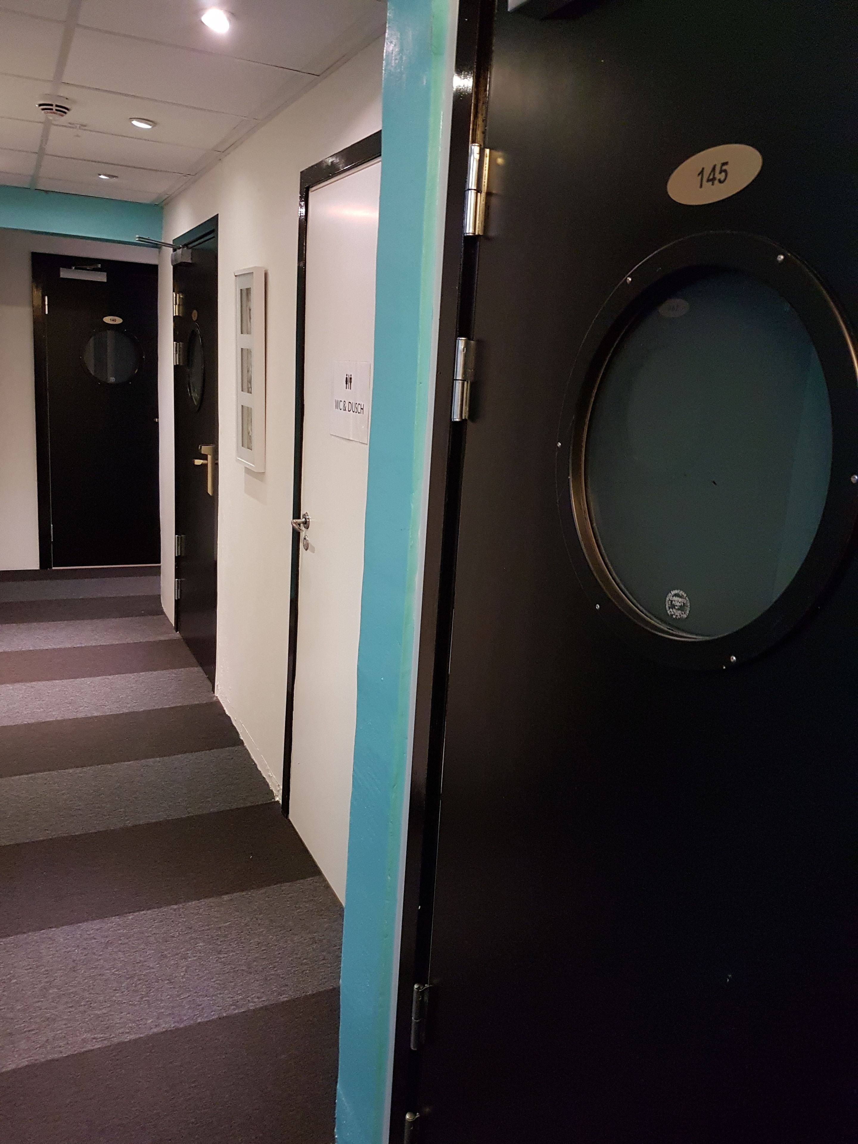 Un pasillo con una puerta negra marcada con el número 145, una pared azul y un cartel que indica WC y ducha.